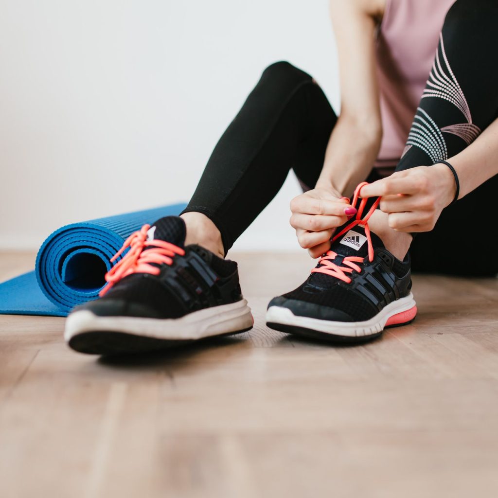 Crop sportswoman in sportswear tying sneakers while sitting on wooden floor with sport mat and dumbbells before exercising in