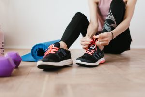 Crop sportswoman in sportswear tying sneakers while sitting on wooden floor with sport mat and dumbbells before exercising in