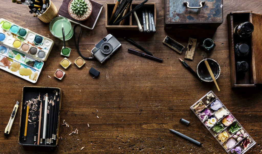 An overhead view of an artist's workspace with watercolor paints, pencils, and various tools.
